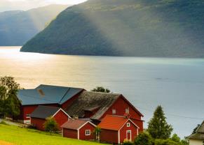 sunrays beam down on a red and white barn house located off the shoreline of a fjord