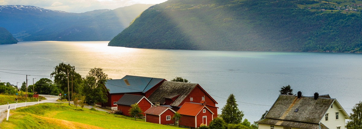 sunrays beam down on a red and white barn house located off the shoreline of a fjord