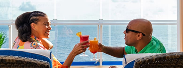 man and woman toast with their tropical drinks while sitting on their private stateroom balcony with ocean view