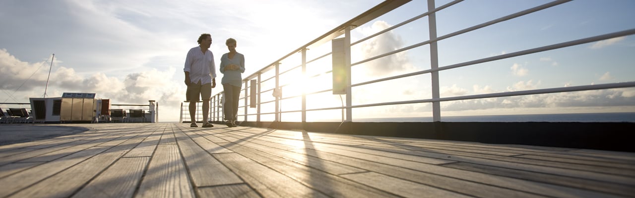 Man and woman walking down wooden deck on a carnival ship as the sun sets behind them