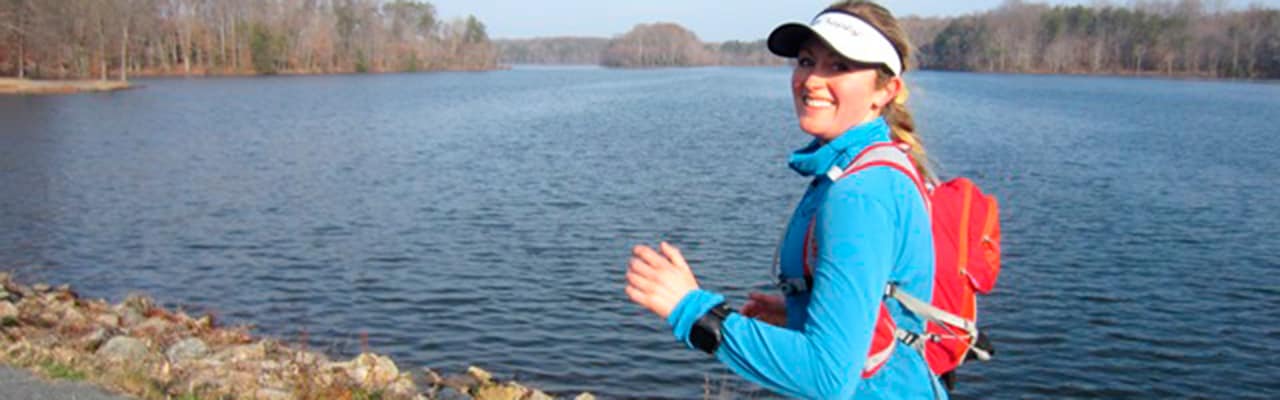 Woman smiles at camera in hiking gear next to a wooded lake