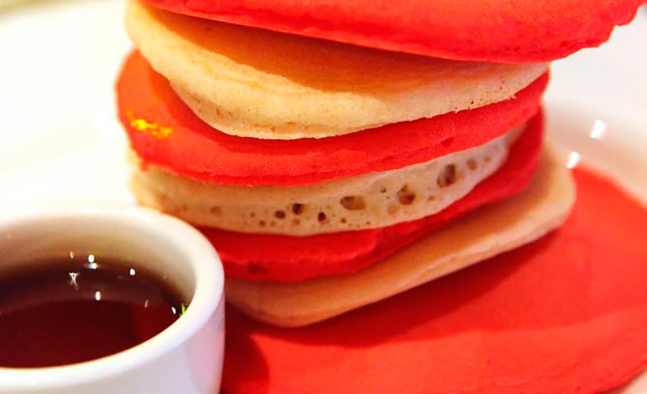A close up of stacked red and white pancakes with a small bowl of syrup