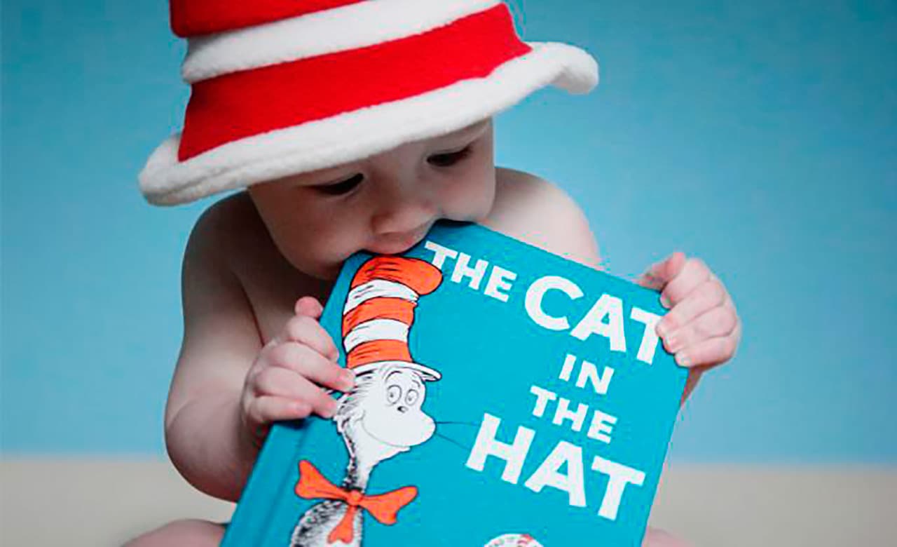 Baby wearing red and white stripe hat chews on corner of "The Cat in the Hat" book