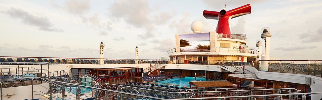 A side view of the ship's deck: pool, beach chairs, lights, TV screen and funnel