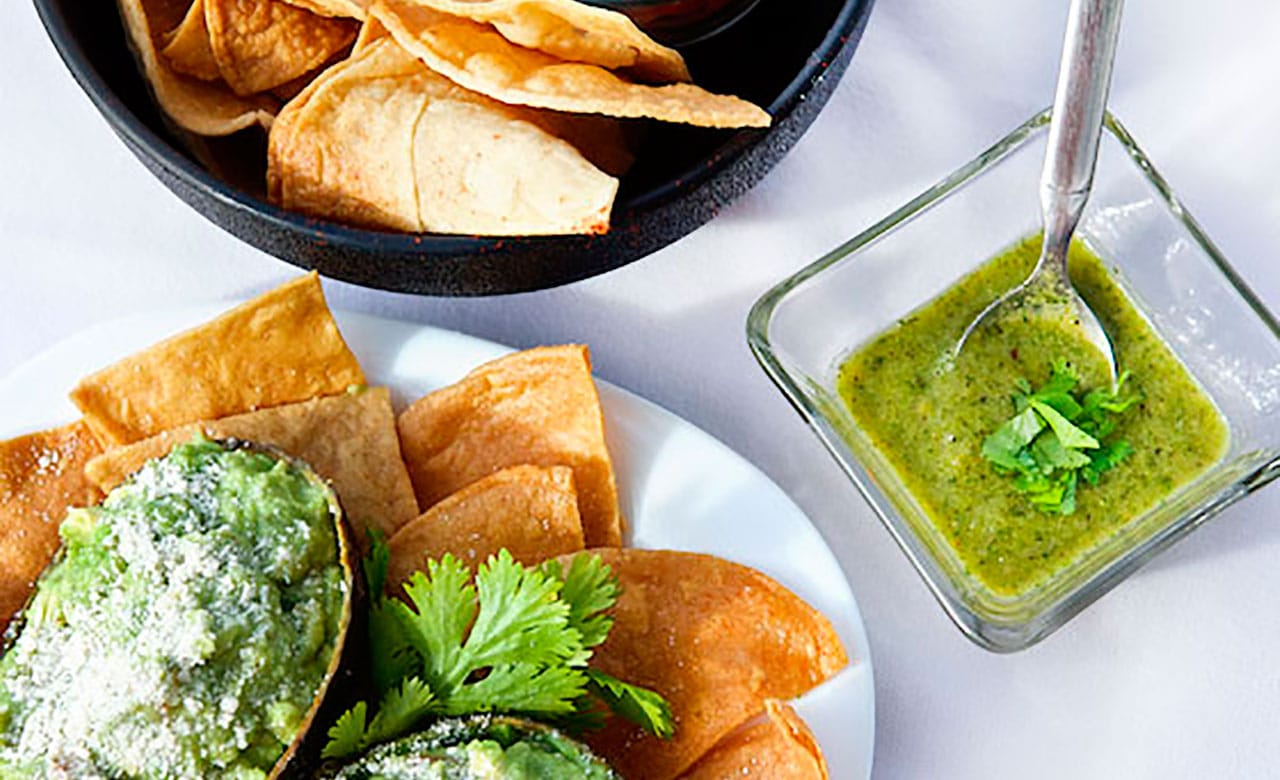 Plated triangular tortilla chips covered in guacamole next to a square glass bowl of green liquid with a spoon placed inside