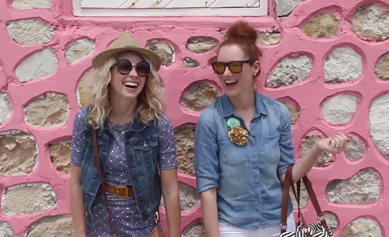 Two women wearing sunglasses, laughing in front of pink and white clay building