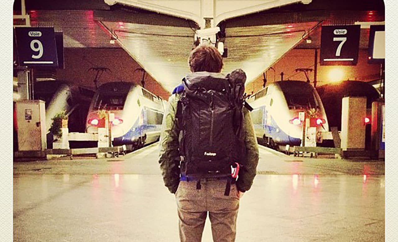 Man stands facing row of trains, wearing a backpack