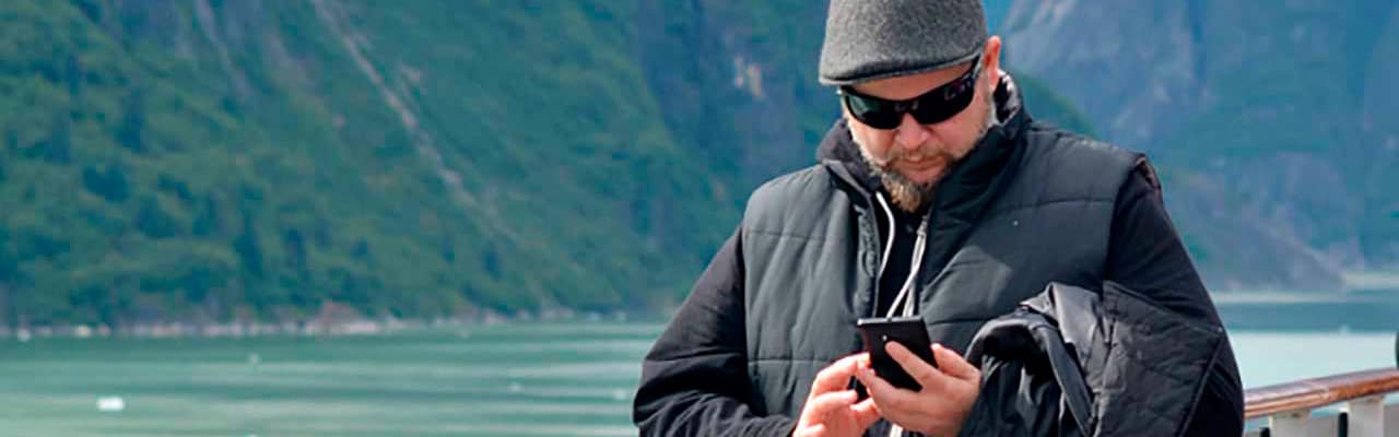 Man wearing heavy vest and winter hat looks at his phone with mountains in the background