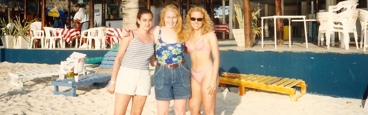 Three girls stand in sand with an outdoor bar behind them