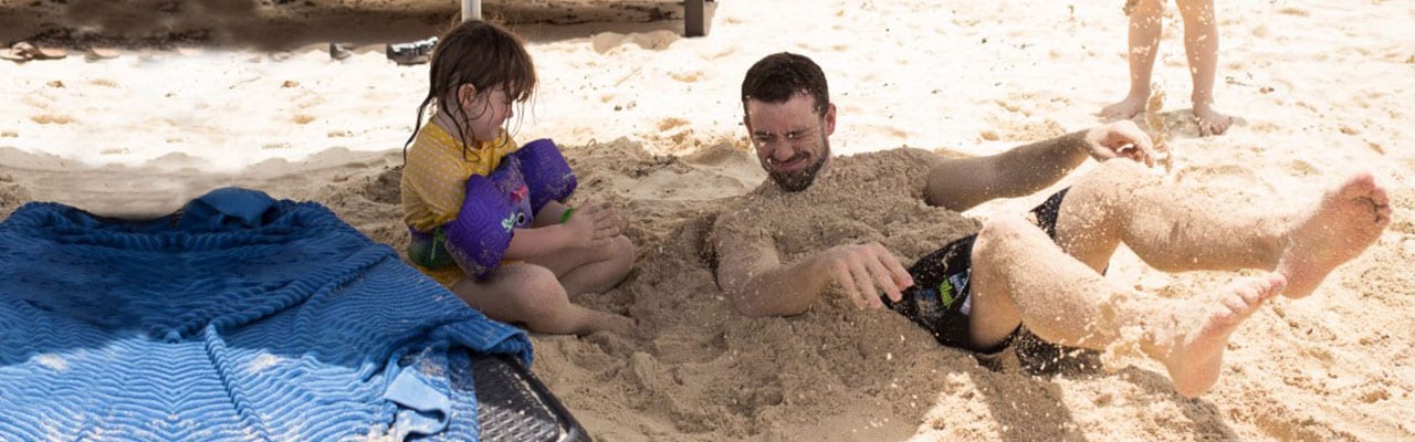 Man, laying and covered in sand on beach as young girl sits beside him