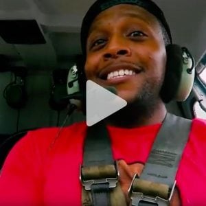 Man with helicopter headphones on, strapped in on a helicopter smiling, link to Youtube video