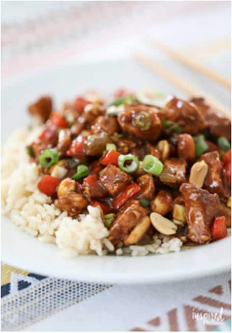 Kung Pao Chicken dish over rice on a white plate sitting on a colorful table cloth