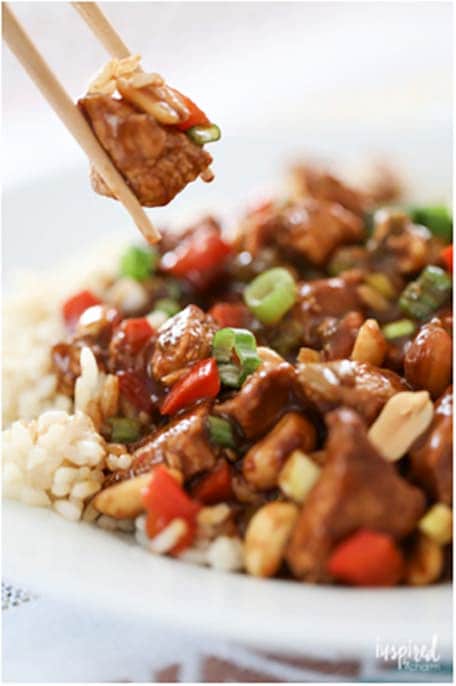 Two chopsticks holding a piece of chicken over a Kung Pao Chicken dish