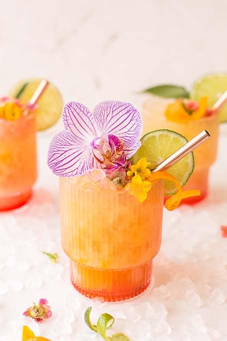 Pineapple mango rum punch cocktail garnished with large pink flower, lime slice and orange zest