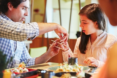 a father teaching his daughter how to use chopsticks to pick up sushi