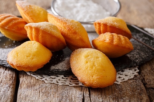 a fresh batch of madeleines on a wooden table with some powdered sugar