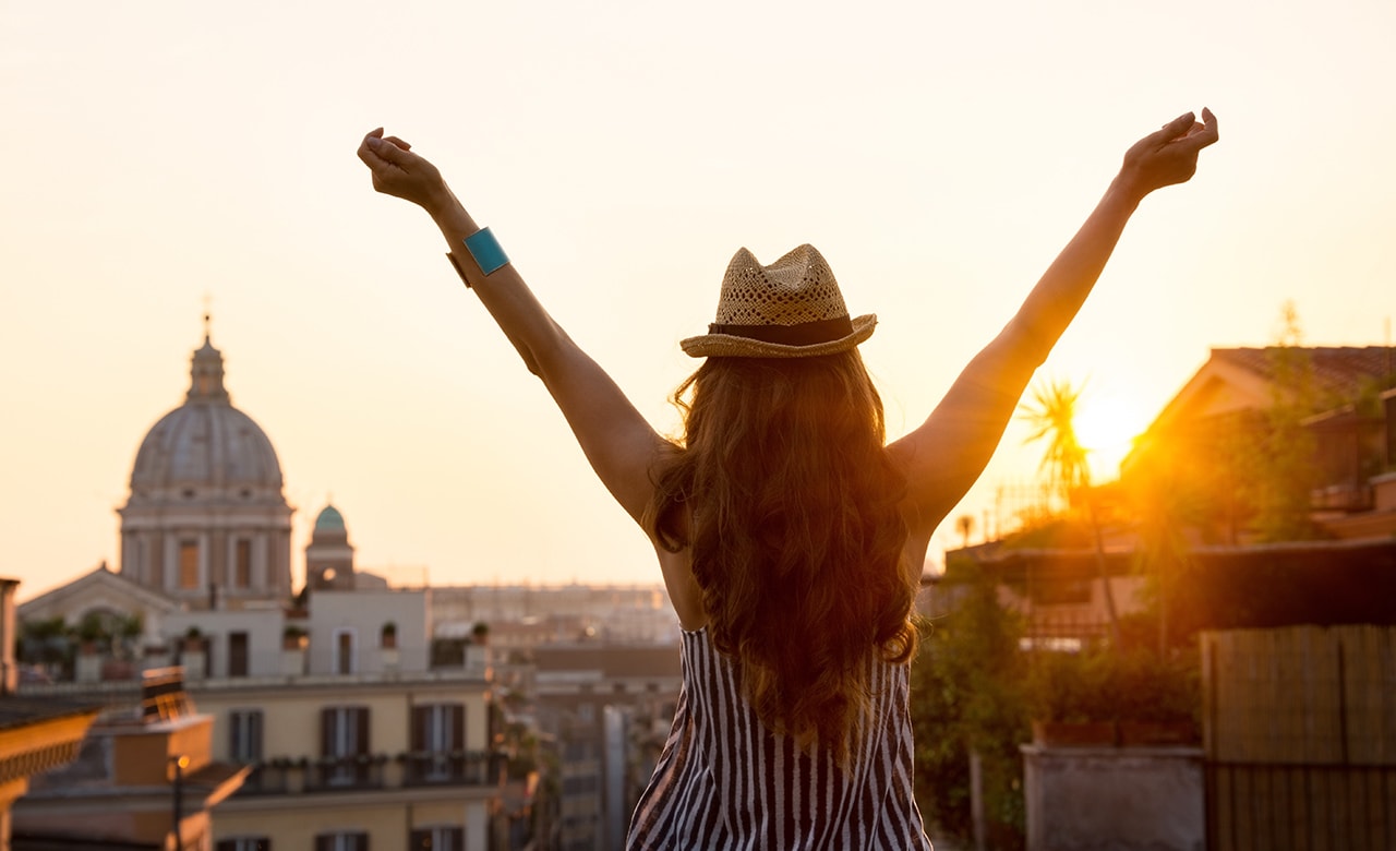 Seen from behind, a woman is standing with outstretched arms, looking out at the city of Rome at sunset in summer.