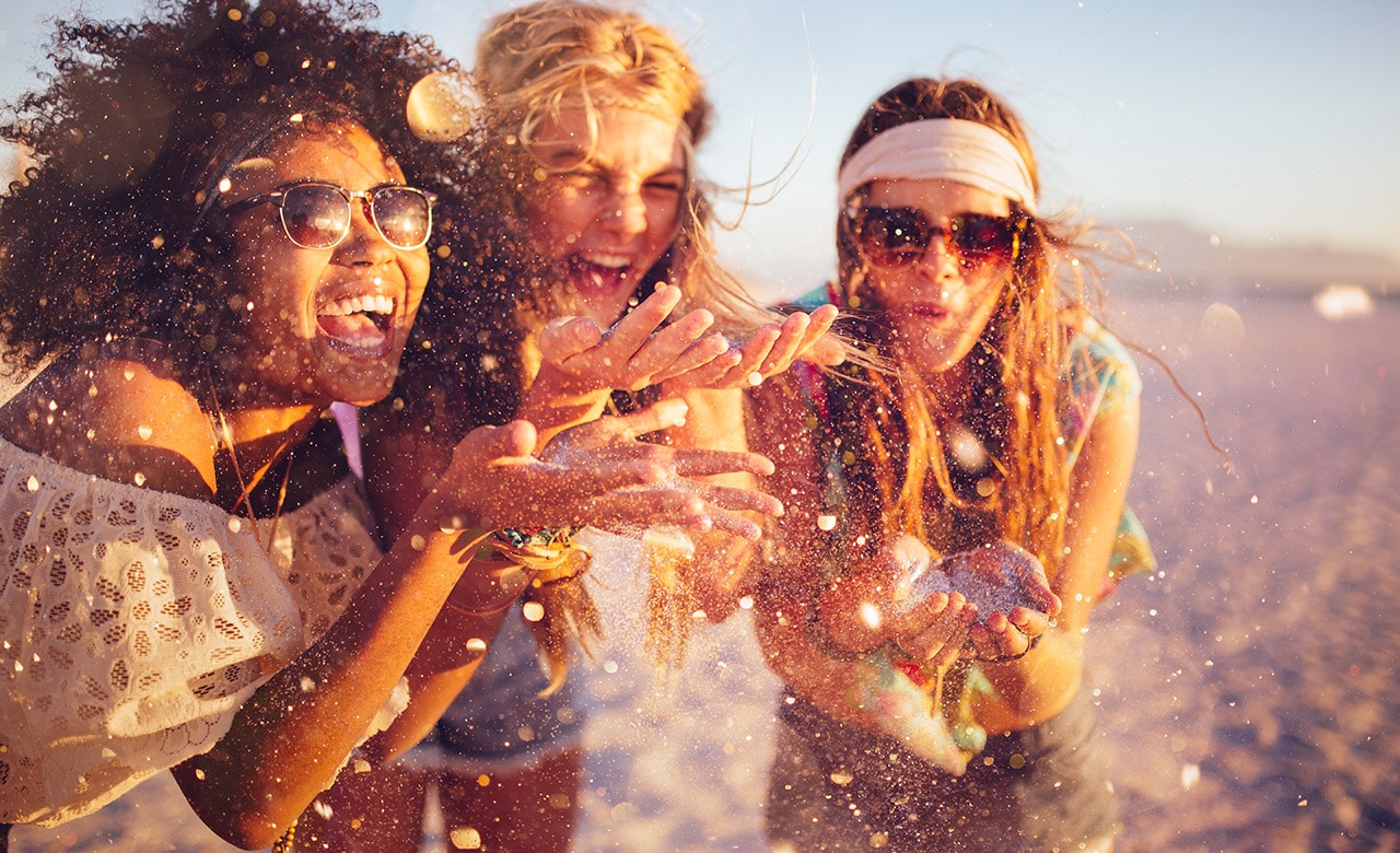 Mixed race group of girls blowing colourful confetti from their hands happily on a beach at sunset