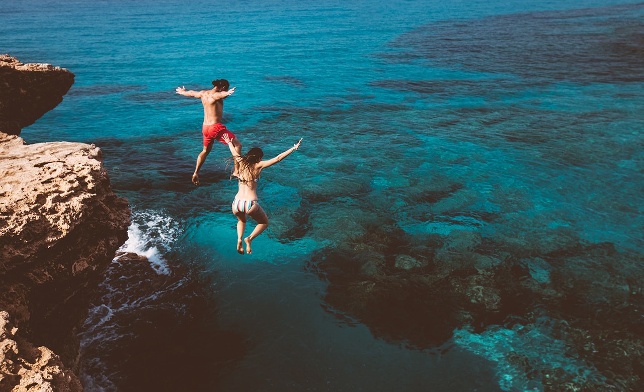 Young active man and woman diving from high cliff into tropical island blue sea water