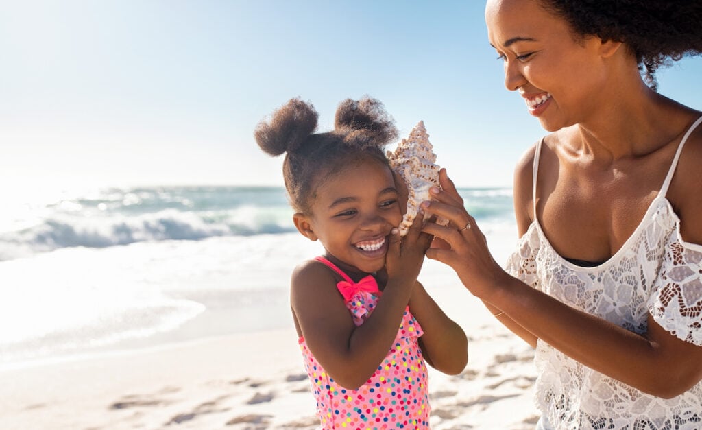 A happy mother holding a seashell to the ear of her daughter at a beautiful beach.