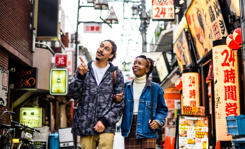 Travelers walking along a city street surrounded by shops with signs in foreign languages.